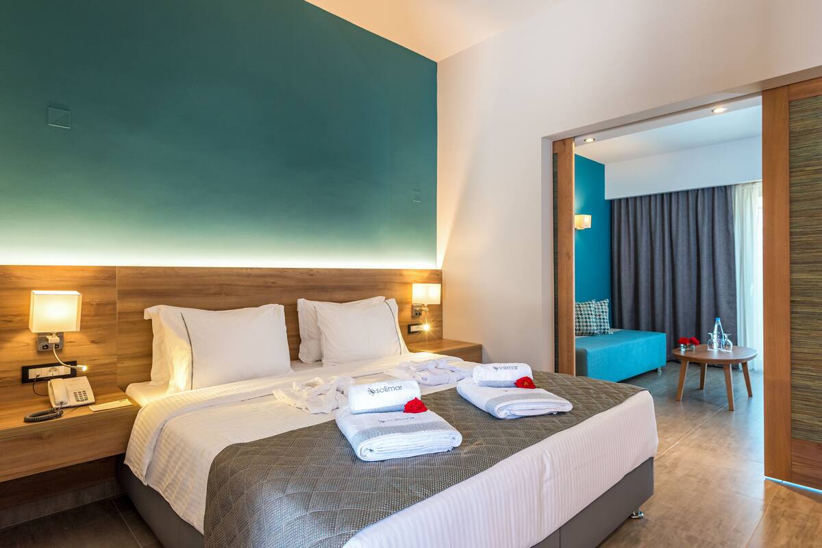 A hotel room with a large bed, two bedside tables with lamps, a wooden headboard, a blue accent wall, and a view of a balcony with curtains.