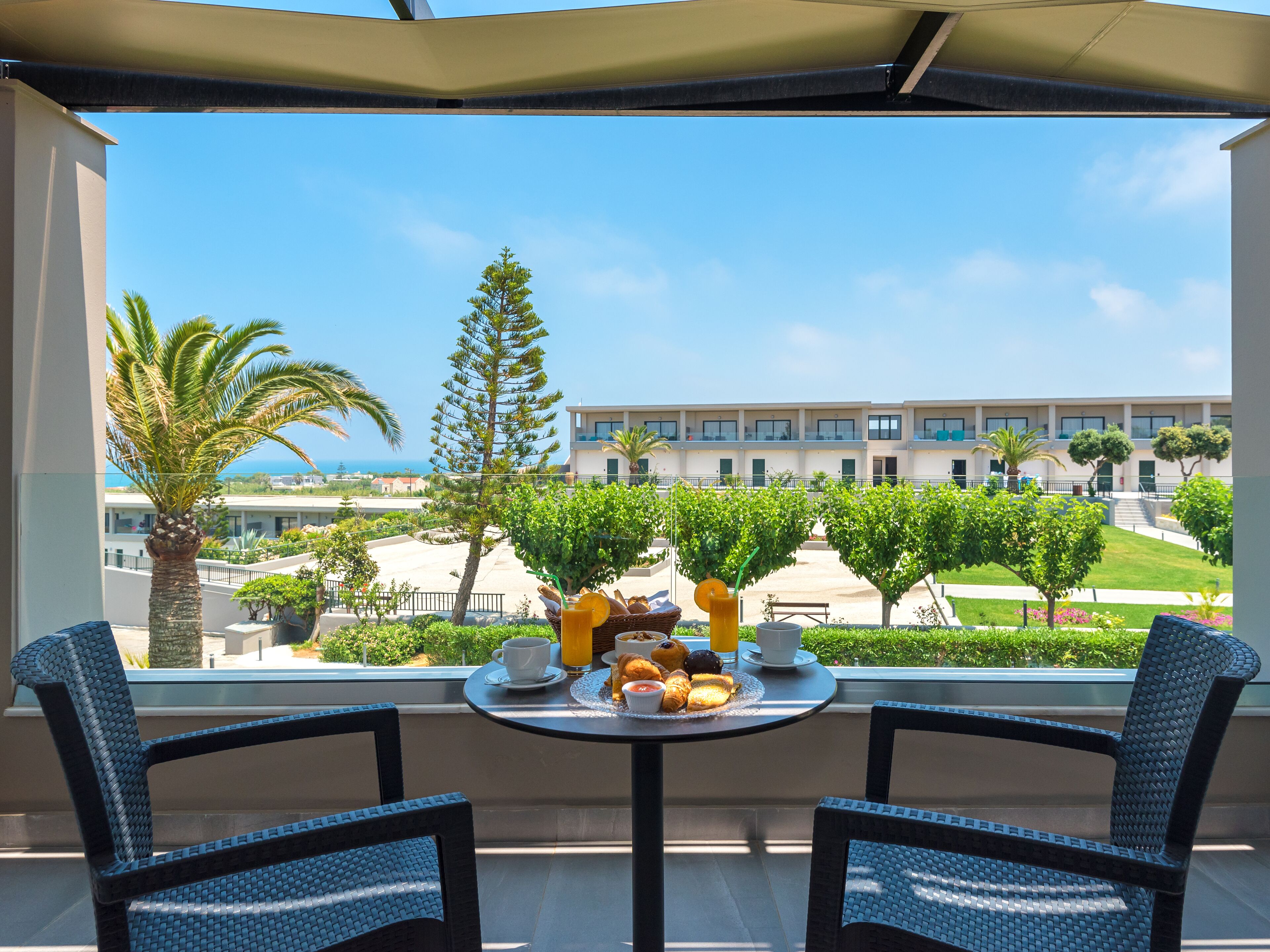 A balcony with two wicker chairs, a table with breakfast items, and a view of a building and greenery.