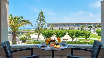 A balcony with two wicker chairs, a table with breakfast items, and a view of a building and greenery.