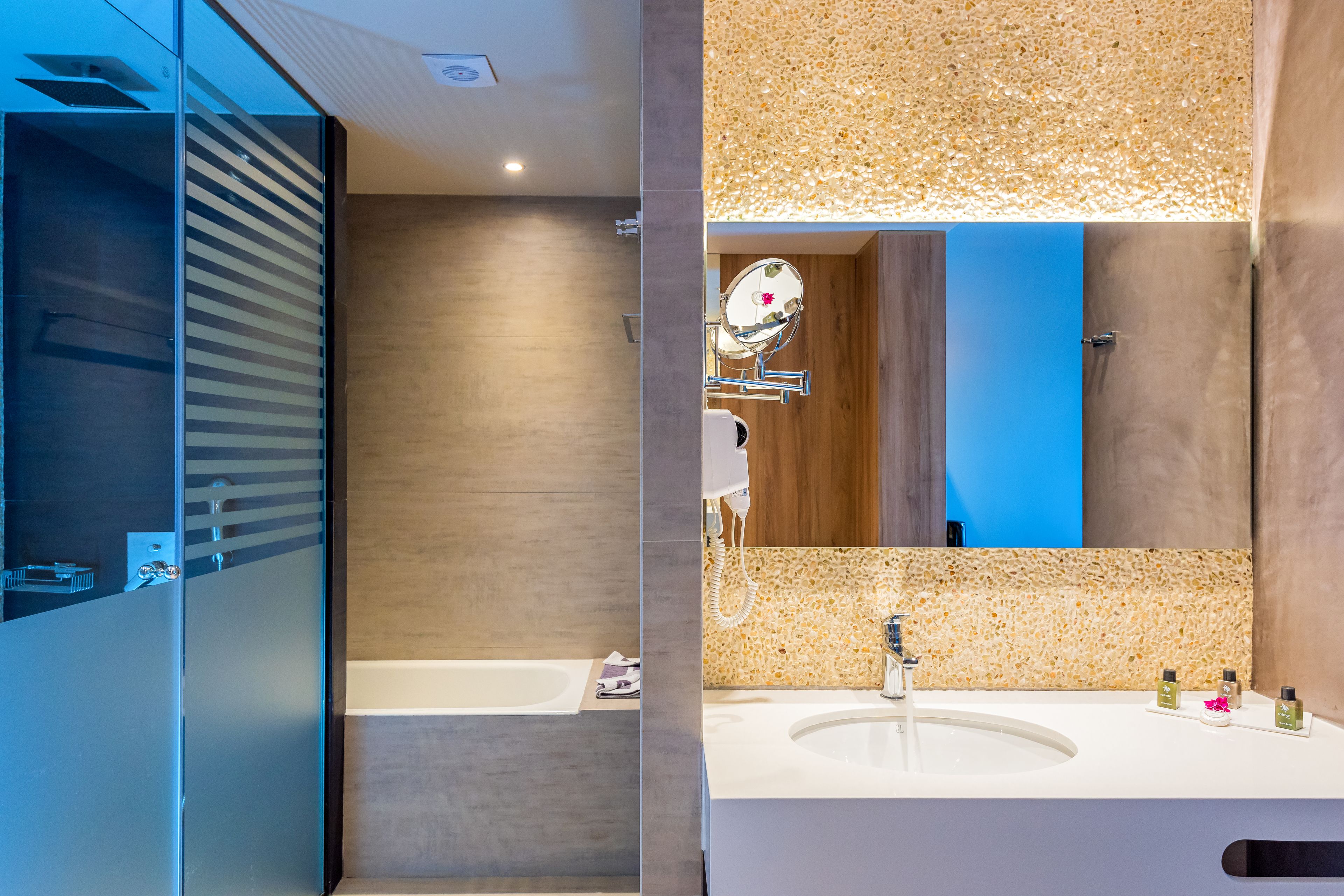 Un baño moderno con separador de vidrio, bañera, lavamanos con artículos de aseo y un espejo grande.
