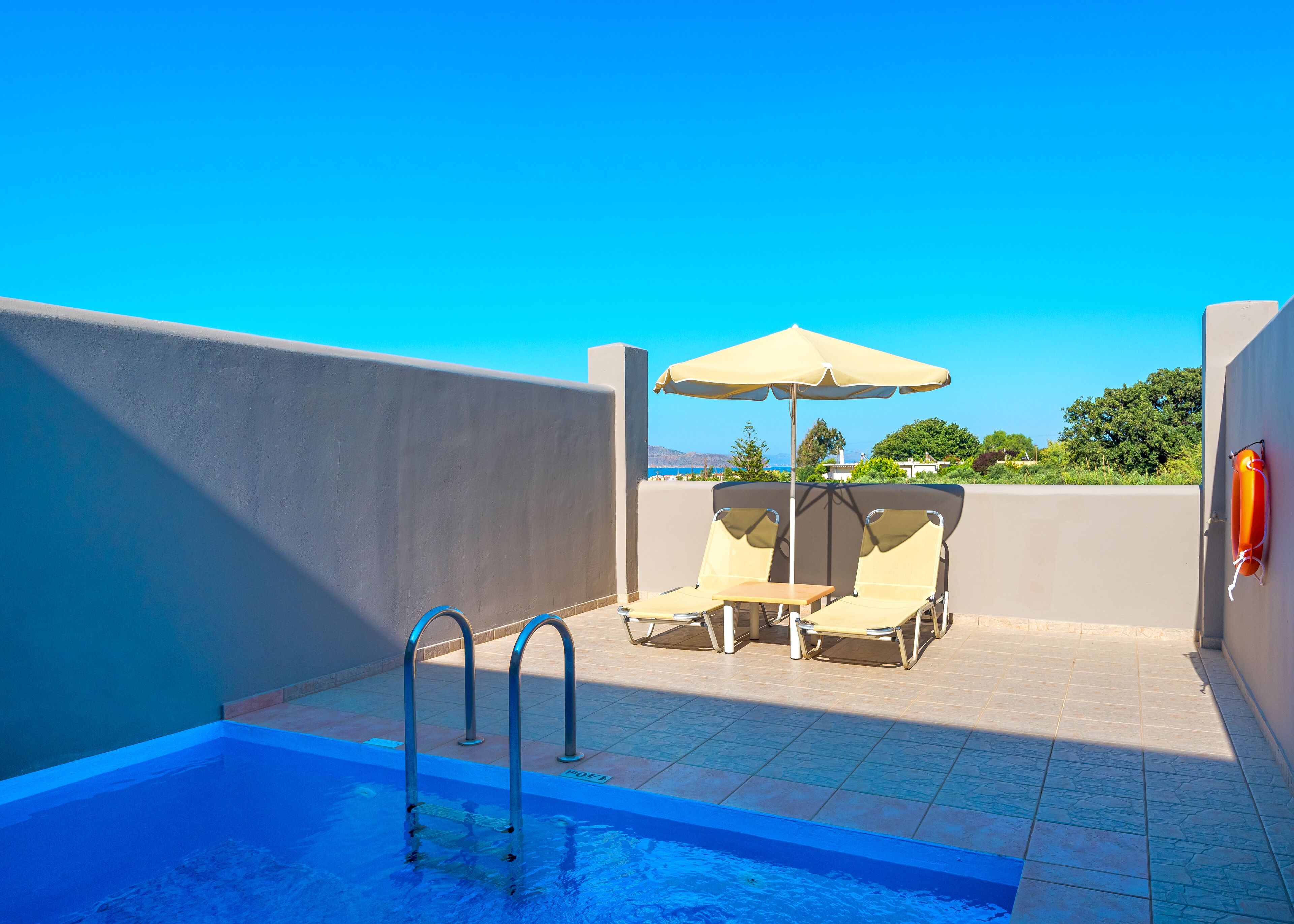 A rooftop pool area with two lounge chairs and a table under a large umbrella, overlooking a clear blue sky and distant landscape.