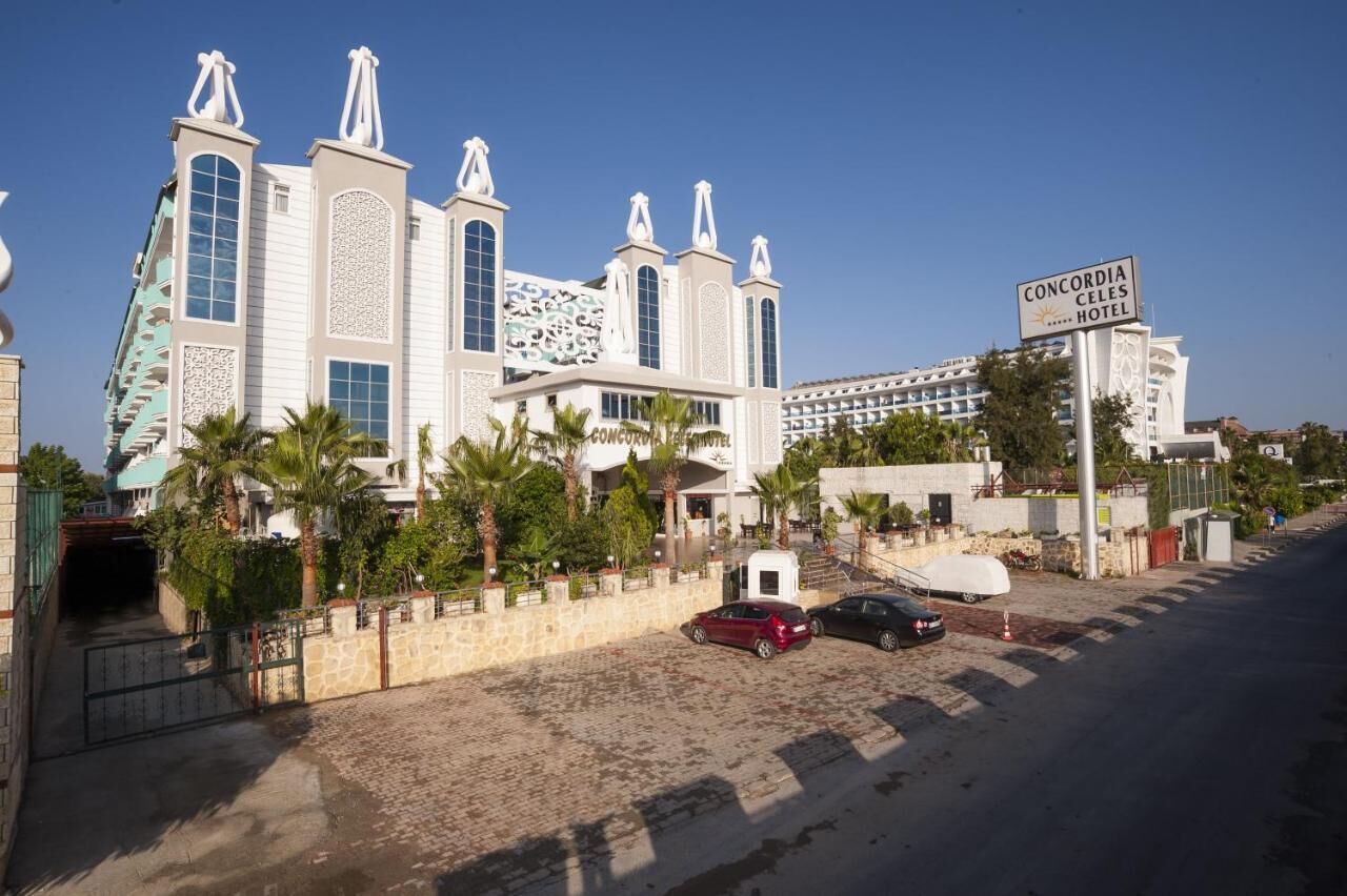 A hotel with a modern architectural design, featuring prominent statues on the facade, a parking area, and a clear sky.