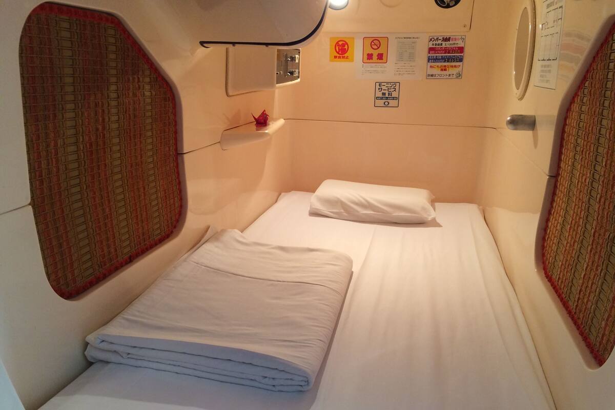 Ein kleines Zimmer mit einem Einzelbett, Kissen und einem kleinen Regal. An der Wand befinden sich Schilder, darunter ein Nichtraucher-Symbol.