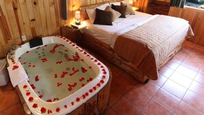 A wooden room with a large bed, a wooden bathtub with rose petals, and a nightstand with a lamp. - Cabañas El Estribo Hotel (Valle de Bravo)