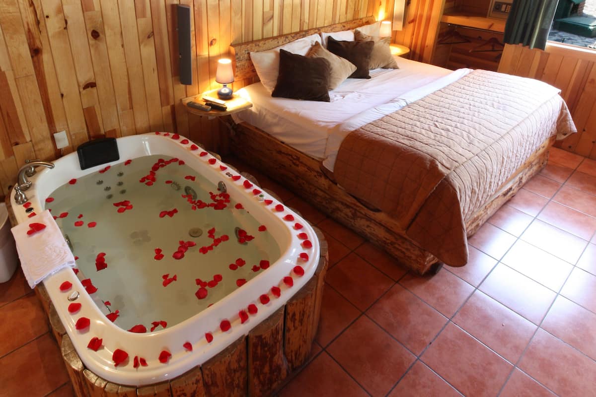A wooden room with a large bed, a wooden bathtub with rose petals, and a nightstand with a lamp.