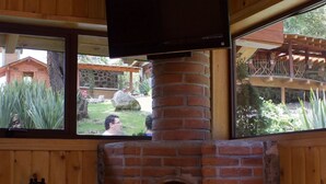 A brick fireplace with a TV mounted above it, a wooden bench with a horse sculpture, and a view of a garden through the windows. - Cabañas El Estribo Hotel (Valle de Bravo)