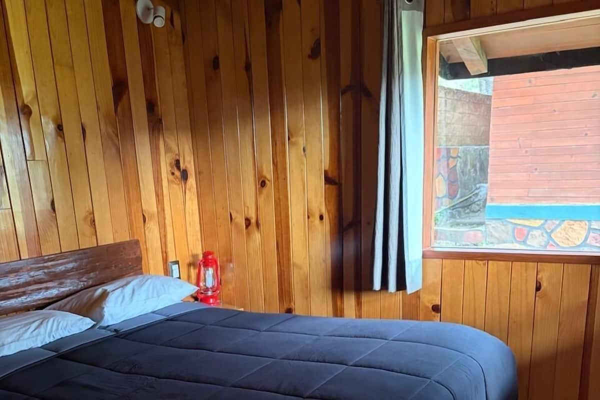 A wooden room with a bed, a window with curtains, and a small table with a bottle.