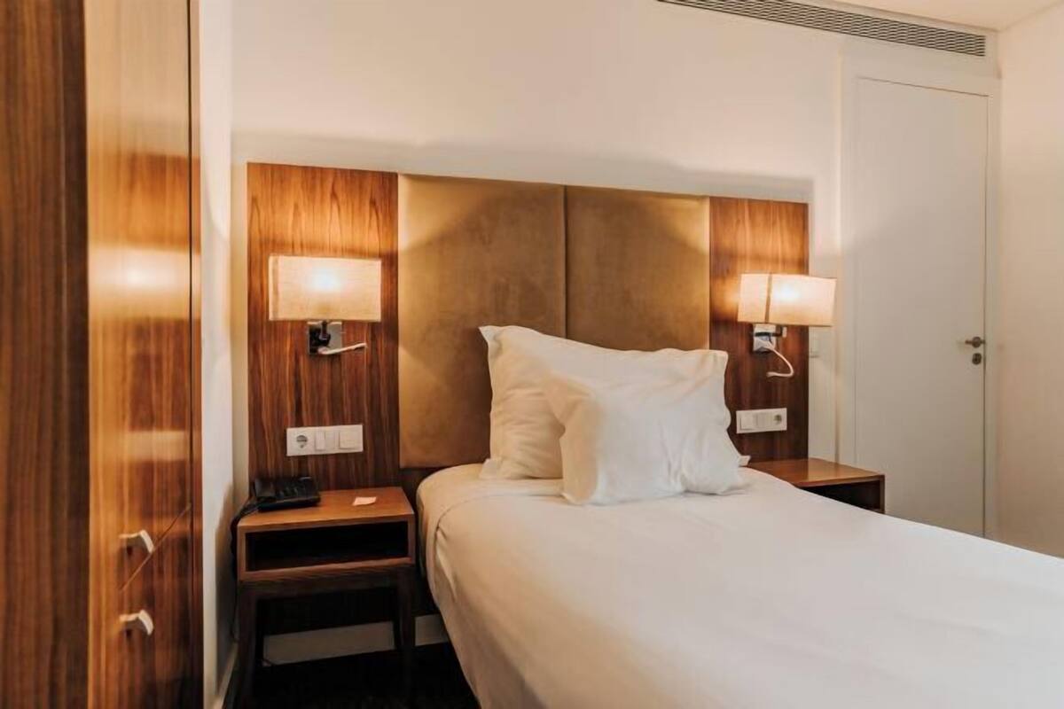 A hotel room with a bed, wooden headboard, bedside lamps, and a nightstand.