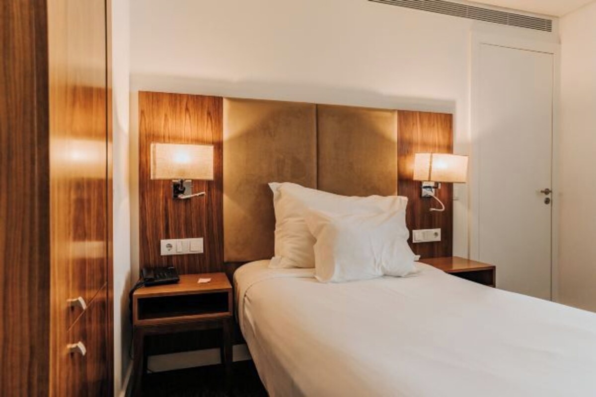 A hotel room with a bed, wooden headboard, bedside lamps, and a nightstand.