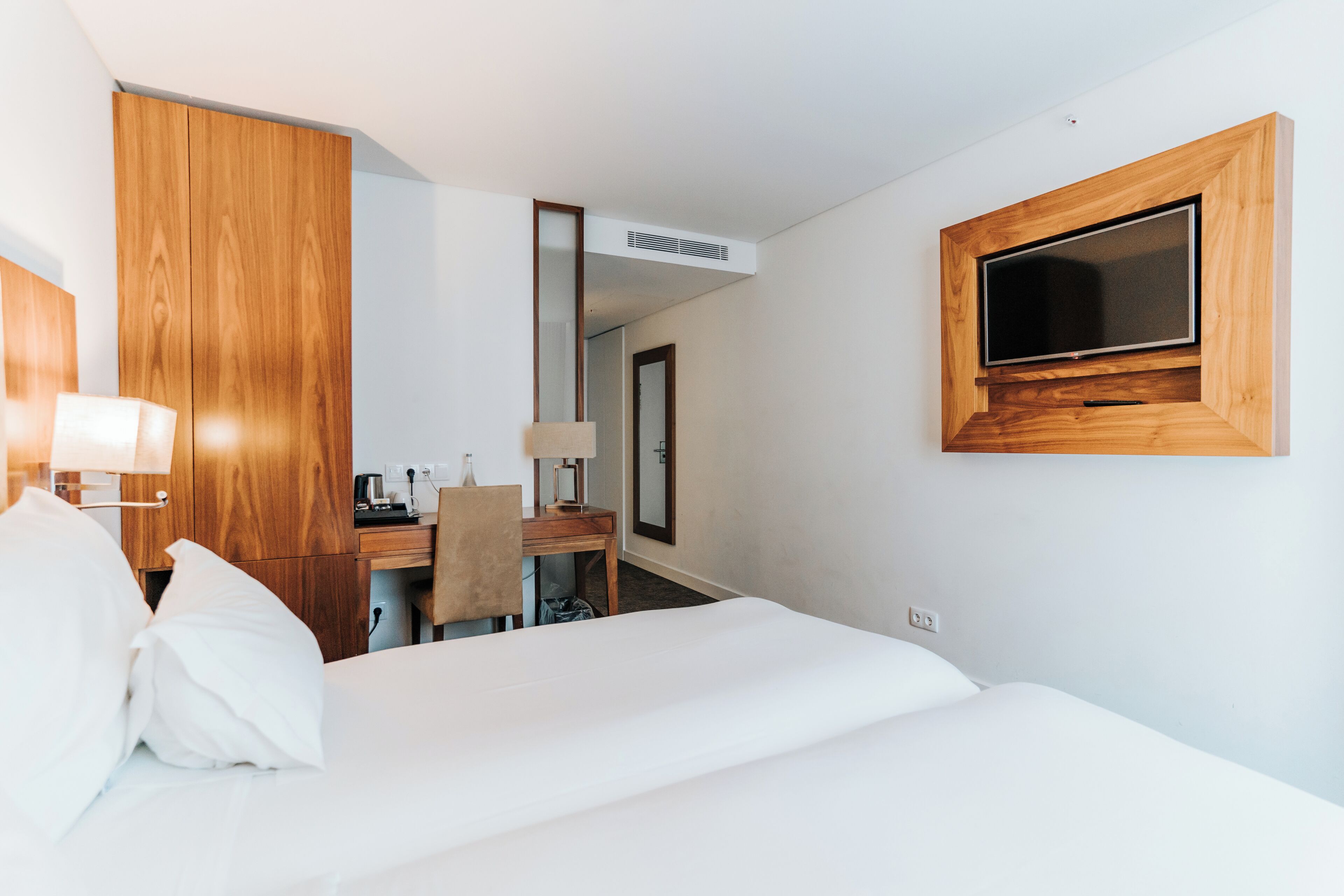 A hotel room with a bed, a desk, a chair, a TV mounted on the wall, and a mirror.