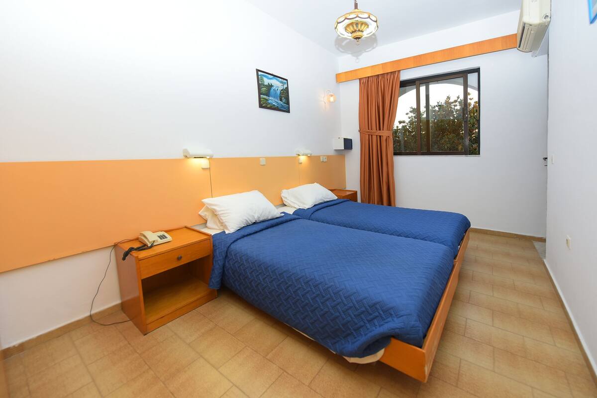 A bedroom with a double bed, a nightstand with a phone, a wall-mounted light, a window with curtains, and a painting on the wall.