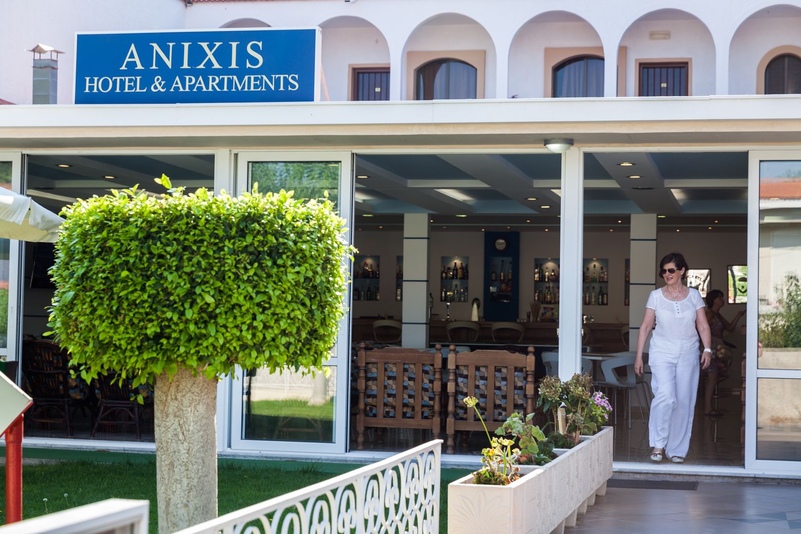 The exterior of Anixis Hotel & Apartments with a large hedge, outdoor seating, and a bar area visible through the glass doors.