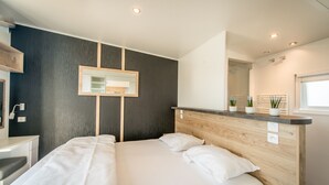 A modern hotel room with a large bed, a wooden headboard, a small bedside table, a mirror, and a window with potted plants. - Holiday Village Knokke (Knokke-Heist)