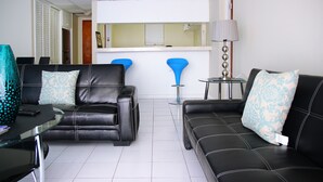 Un salon moderne avec deux canapés en cuir noir, une table basse en verre et une petite table ronde avec une lampe. Au fond, on aperçoit un coin cuisine avec un comptoir blanc et des tabourets de bar bleus.