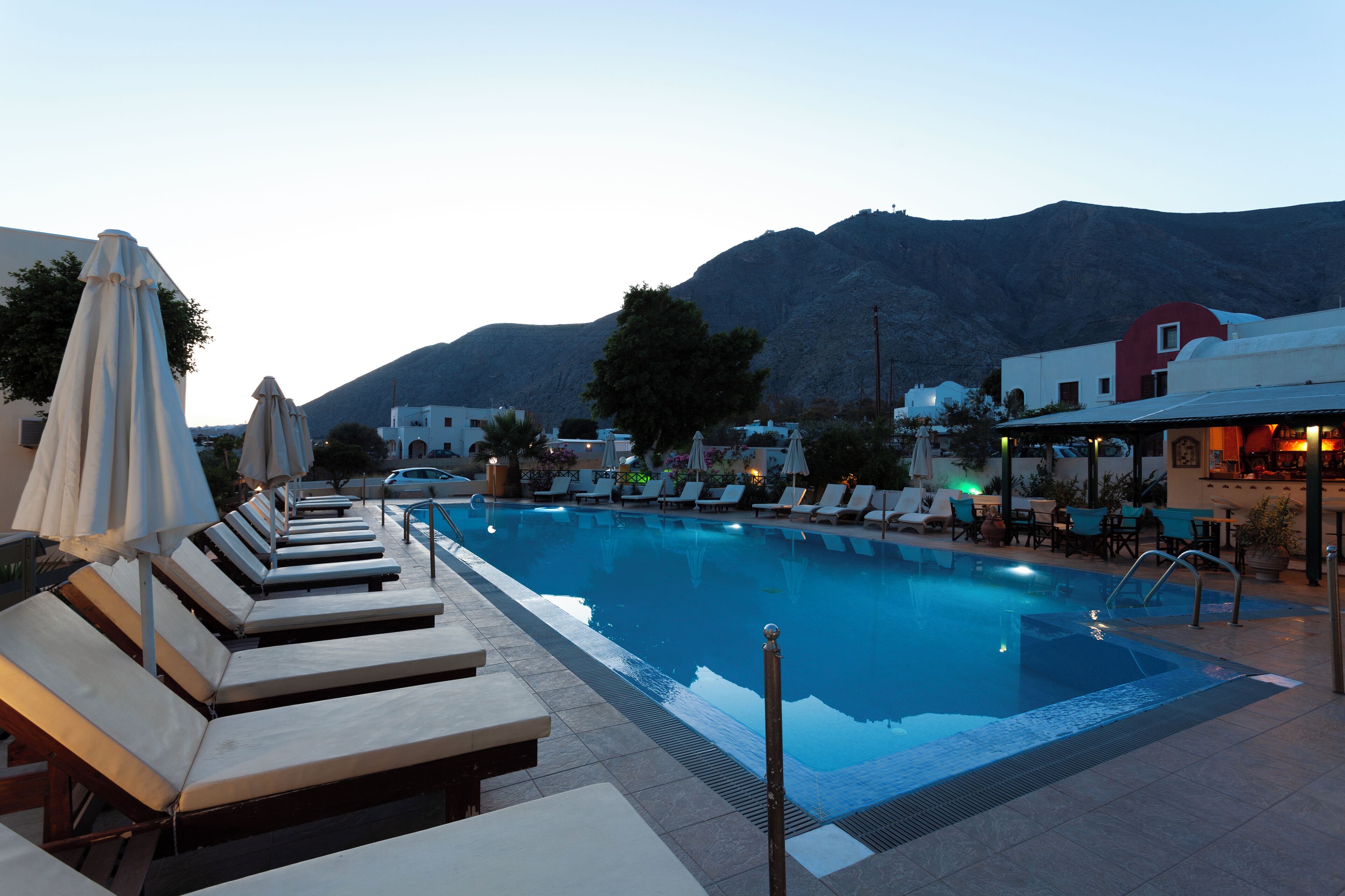 A pool area with lounge chairs and umbrellas, surrounded by buildings and a mountain in the background.