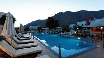 A pool area with lounge chairs and umbrellas, surrounded by buildings and a mountain in the background.