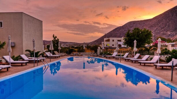 A swimming pool with lounge chairs and umbrellas, surrounded by buildings and a mountain in the background.