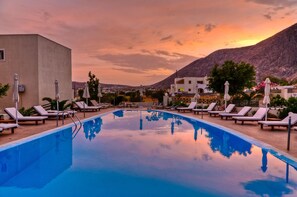 A swimming pool with lounge chairs and umbrellas, surrounded by buildings and a mountain in the background. - Phevos Villa (Santorini)