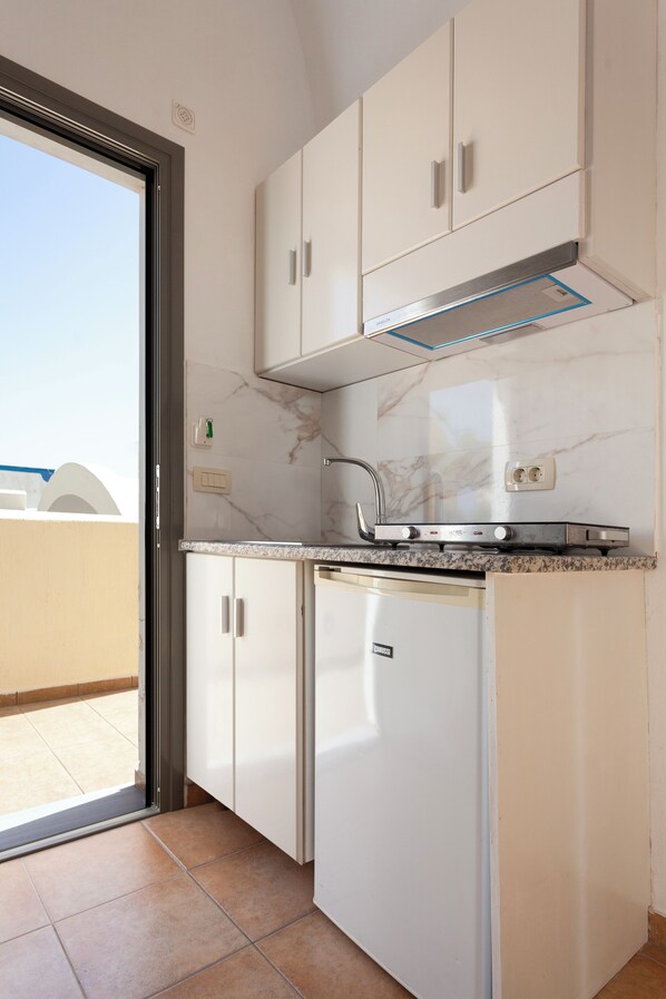 A compact kitchen with white cabinets, a built-in stove, and a window for natural light. - Phevos Villa (Santorini)