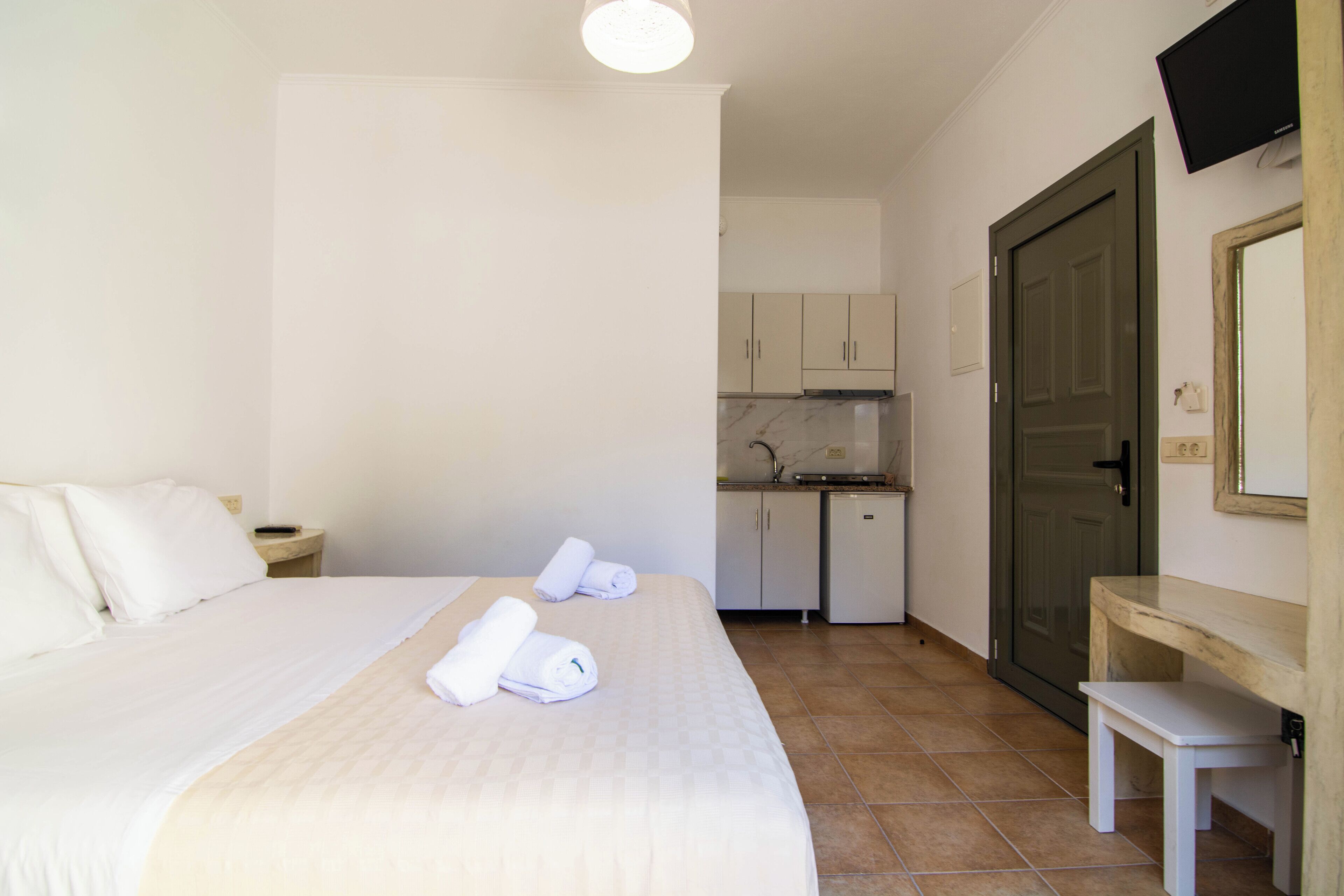 A hotel room with a bed, a small table, a desk, a kitchenette, and a door.