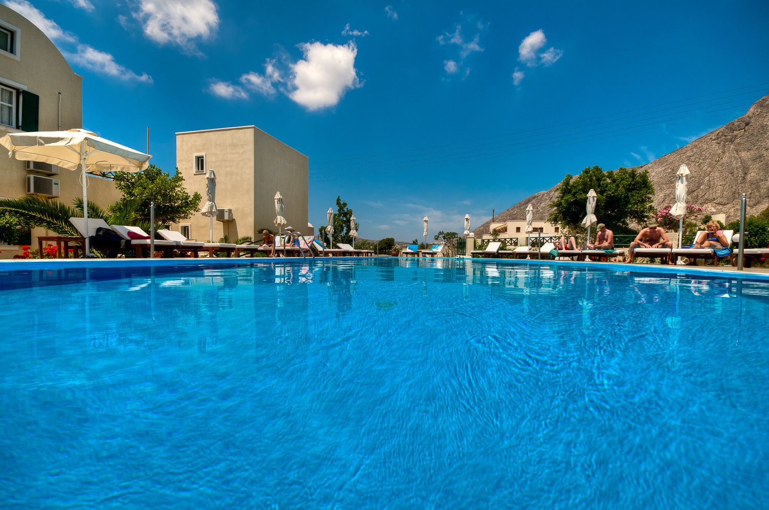 A swimming pool with lounge chairs and umbrellas, surrounded by buildings and a clear sky.