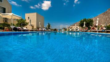A swimming pool with lounge chairs and umbrellas, surrounded by buildings and a clear sky.