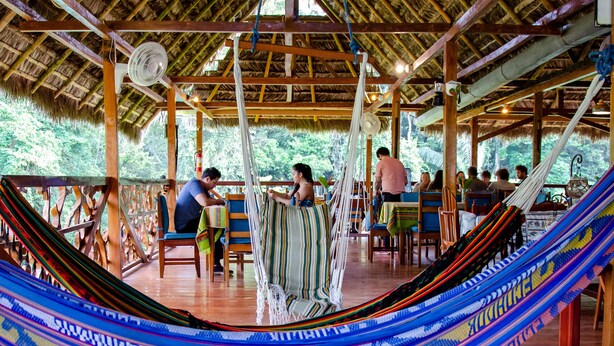 A hammock in a wooden structure with a thatched roof, tables, and chairs.