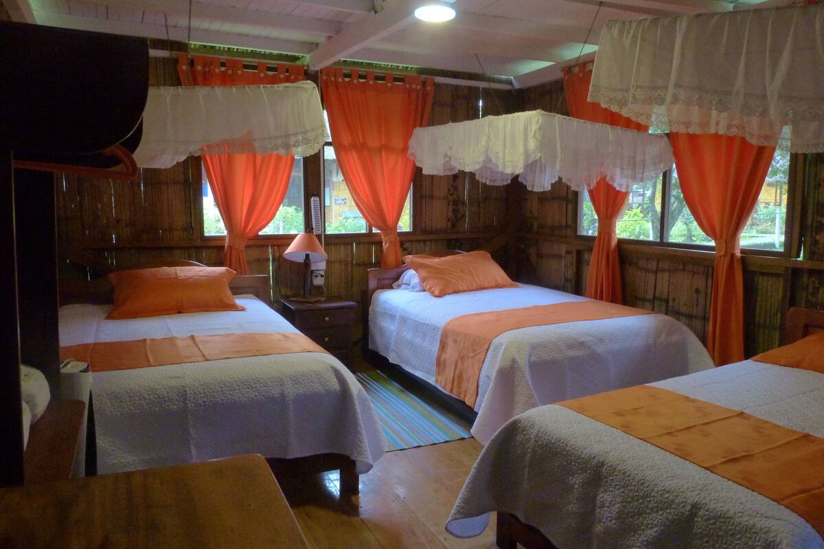 Una habitación con dos camas, cada una con sábanas blancas y almohadas naranjas, y cortinas color naranja.