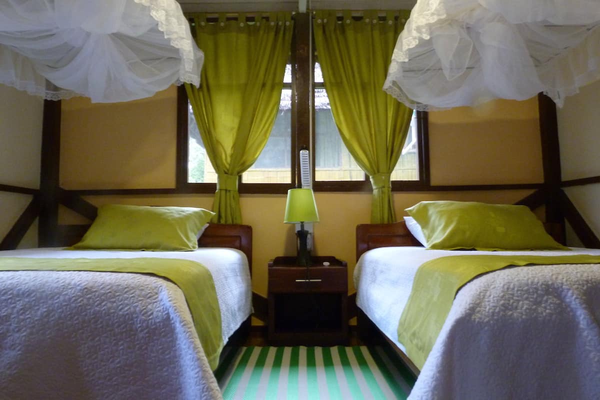 Habitación de hotel con dos camas, cortinas verdes, una mesita de noche con lámpara y piso a rayas.