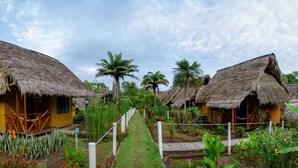 A row of thatched-roof huts with wooden walls, a central pathway, and tropical vegetation.