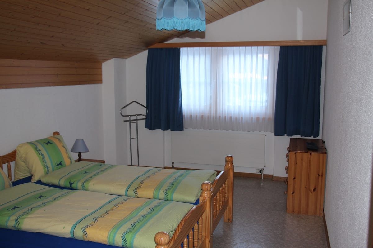 Una habitación con dos camas de madera, un armario pequeño de madera, una lámpara de pie y una ventana con cortinas.