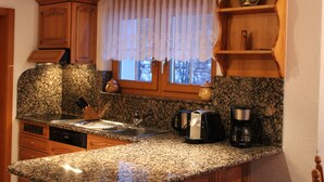 A kitchen with granite countertops, wooden cabinets, a window with a sheer curtain, a coffee maker, and a kettle. - Apartments Arcadia (Saas Grund)