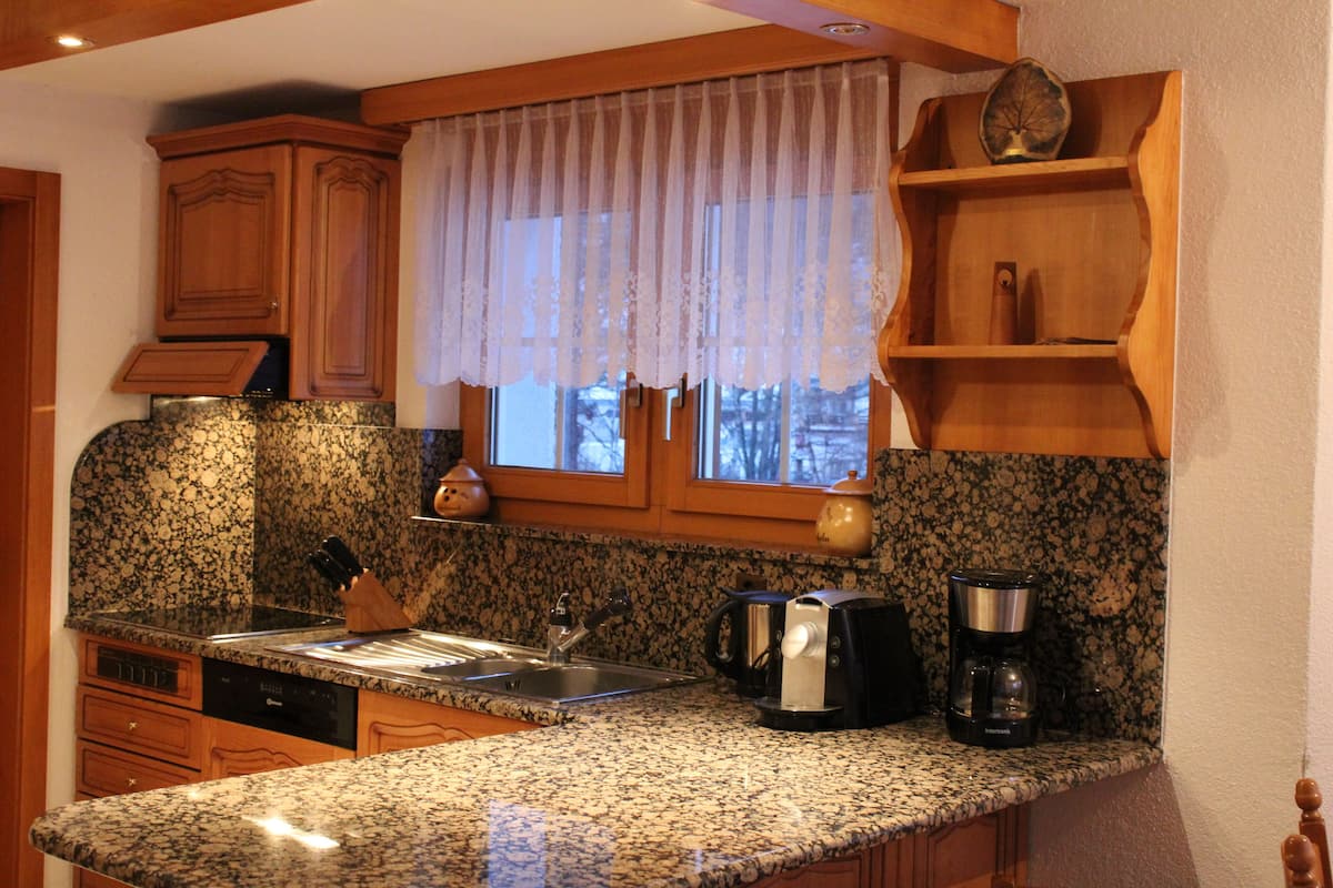 Una cocina con encimeras de granito, armarios de madera, un ventanal con cortina ligera, una cafetera y un hervidor.