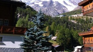 A mountain lodge with a view of snow-capped peaks, surrounded by greenery and a wooden deck. - Apartments Arcadia (Saas Grund)