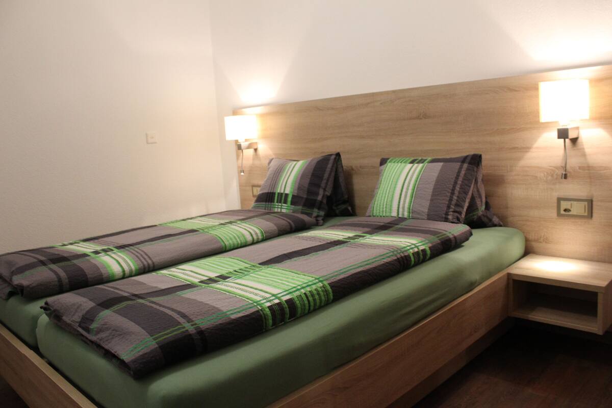 Una cama individual con colcha verde y de cuadros, dos mesitas de noche fijadas a la pared, y un cabecero de madera.
