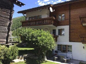 A two-story wooden house with a balcony, a stone bench, and a small table in the yard.