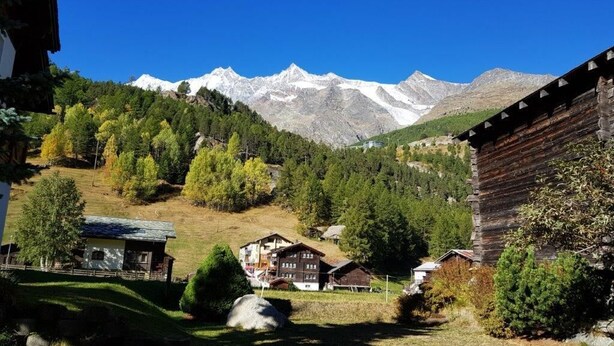 A mountain village with traditional wooden houses, a clear blue sky, and snow-capped peaks in the background. - Apartments Arcadia (Saas Grund)