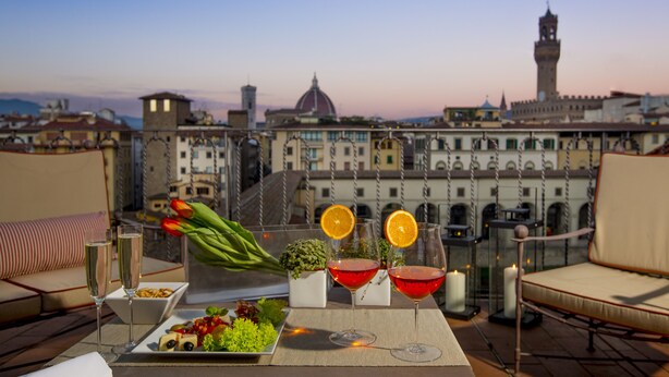 A terrace with a table set for two, featuring a plate of appetizers, two glasses of wine, a vase with tulips, and a view of historic buildings. - Torre Mannelli Suites (Florence)