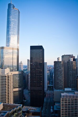 A cityscape with high-rise buildings, including a prominent skyscraper with a unique design and a shorter, rectangular building with a dark facade. - The Penthouse at Grand Plaza (Chicago)