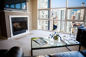 A living room with a fireplace, a glass coffee table with magazines, and a view of high-rise buildings. - The Penthouse at Grand Plaza (Chicago)