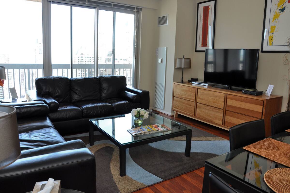 A modern living room with a leather sofa, a glass coffee table, a flat-screen TV, a wooden cabinet, and a large window offering a city view.