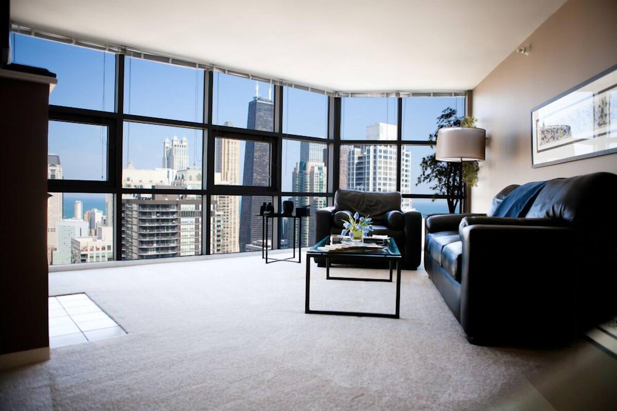 A modern living room with a city view, a black leather sofa, a glass coffee table, and a floor lamp.