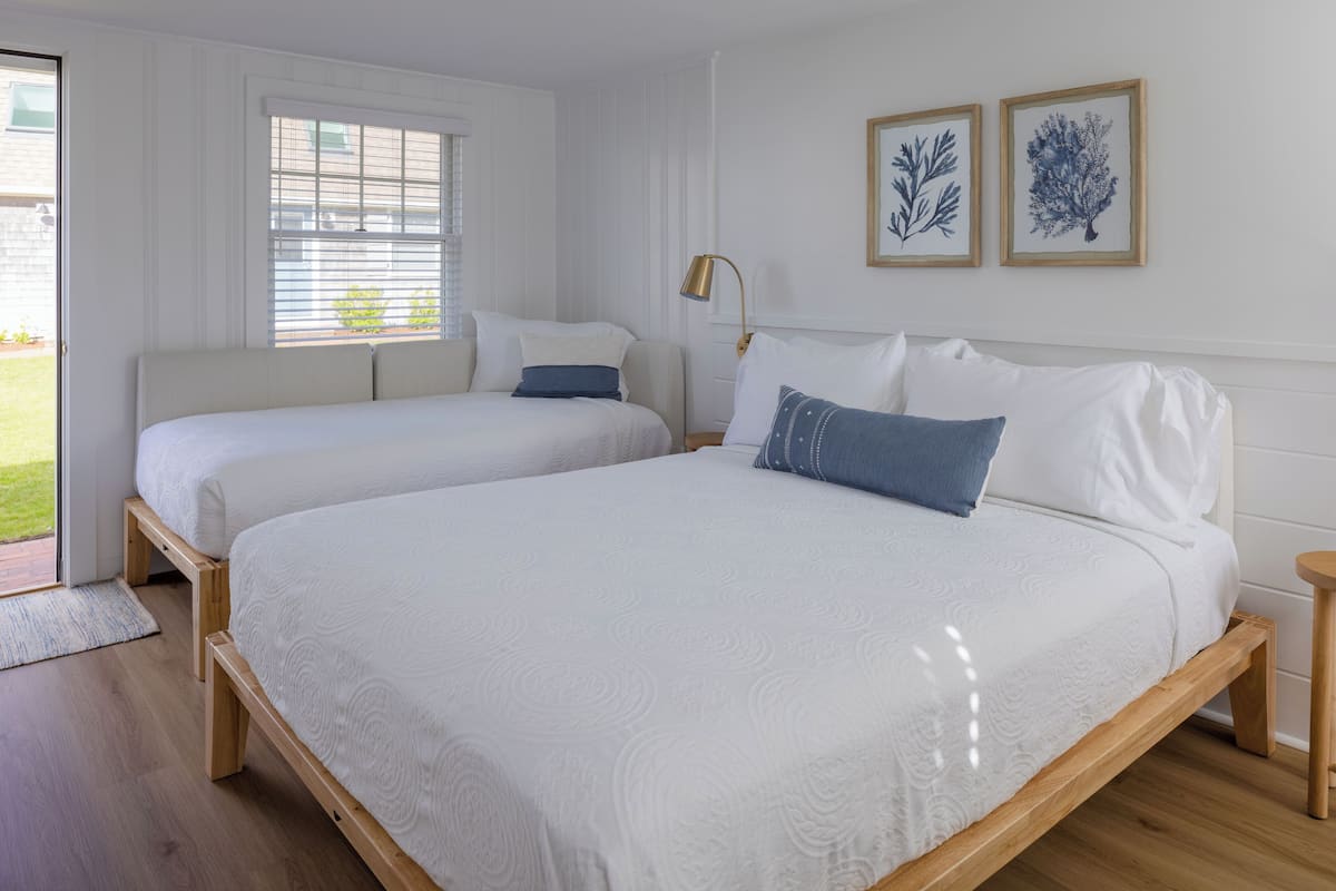 A bedroom with two beds, a wooden floor, a window with blinds, and framed artwork on the wall.