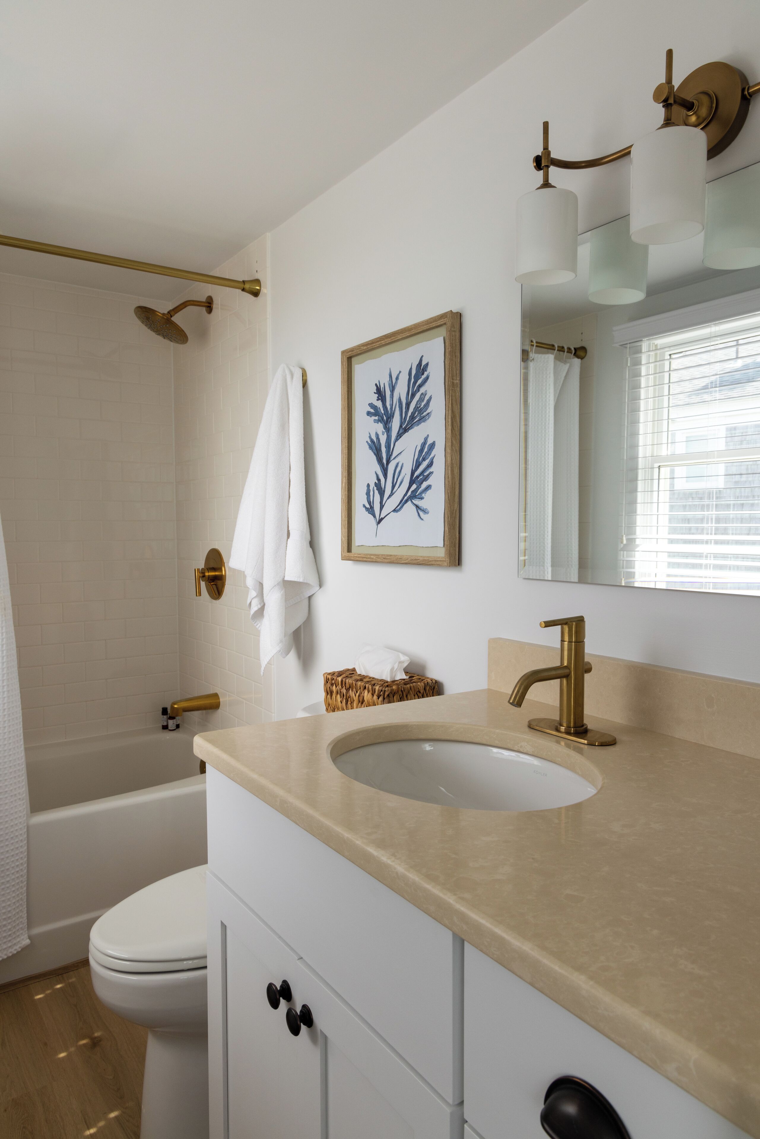 Ein Badezimmer mit Toilette in Weiß, einem beige farbenen Waschbecken mit Goldarmatur, einer Dusche mit goldfarbener Duschbrause und einem gerahmten Kunstwerk an der Wand.