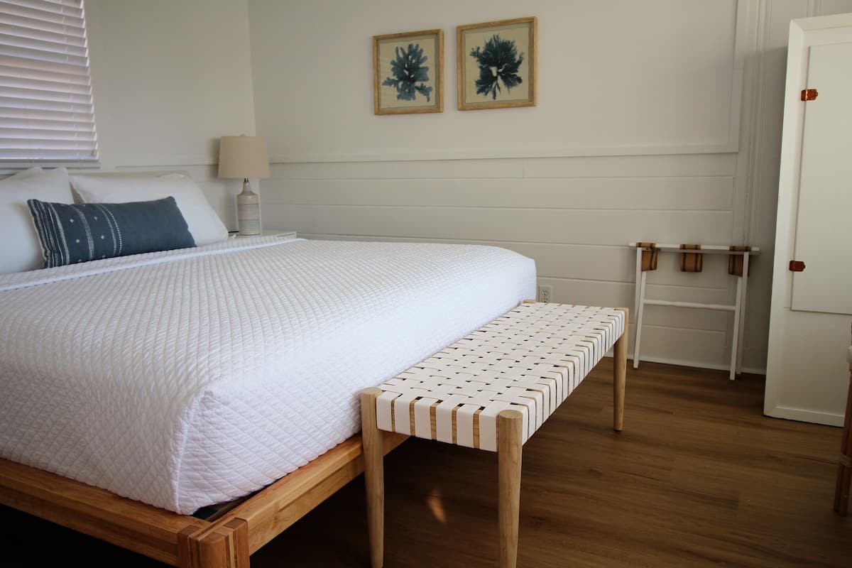 A neatly made bed with a white quilt, a wooden bench, a bedside table with a lamp, and two framed pictures on the wall.