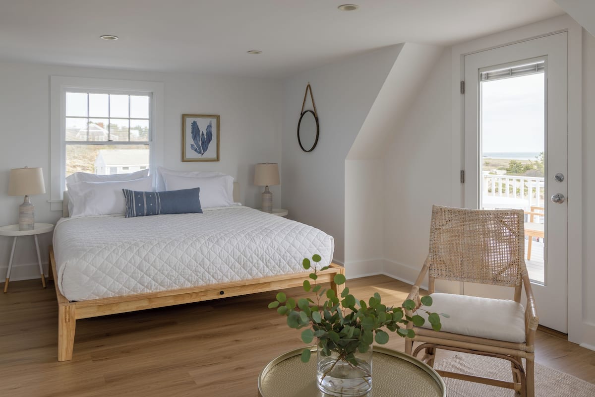 A bedroom with a wooden bed, a wicker chair, a vase with green leaves, and a view of the sea through a door.