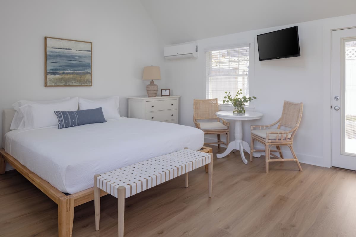 A bedroom with a large bed, a bedside table with a lamp, a chair, a small table with a plant, and a TV mounted on the wall.