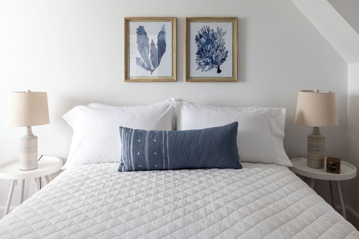 A neatly made bed with a quilted white duvet, two framed botanical prints on the wall, and two bedside lamps on white side tables.