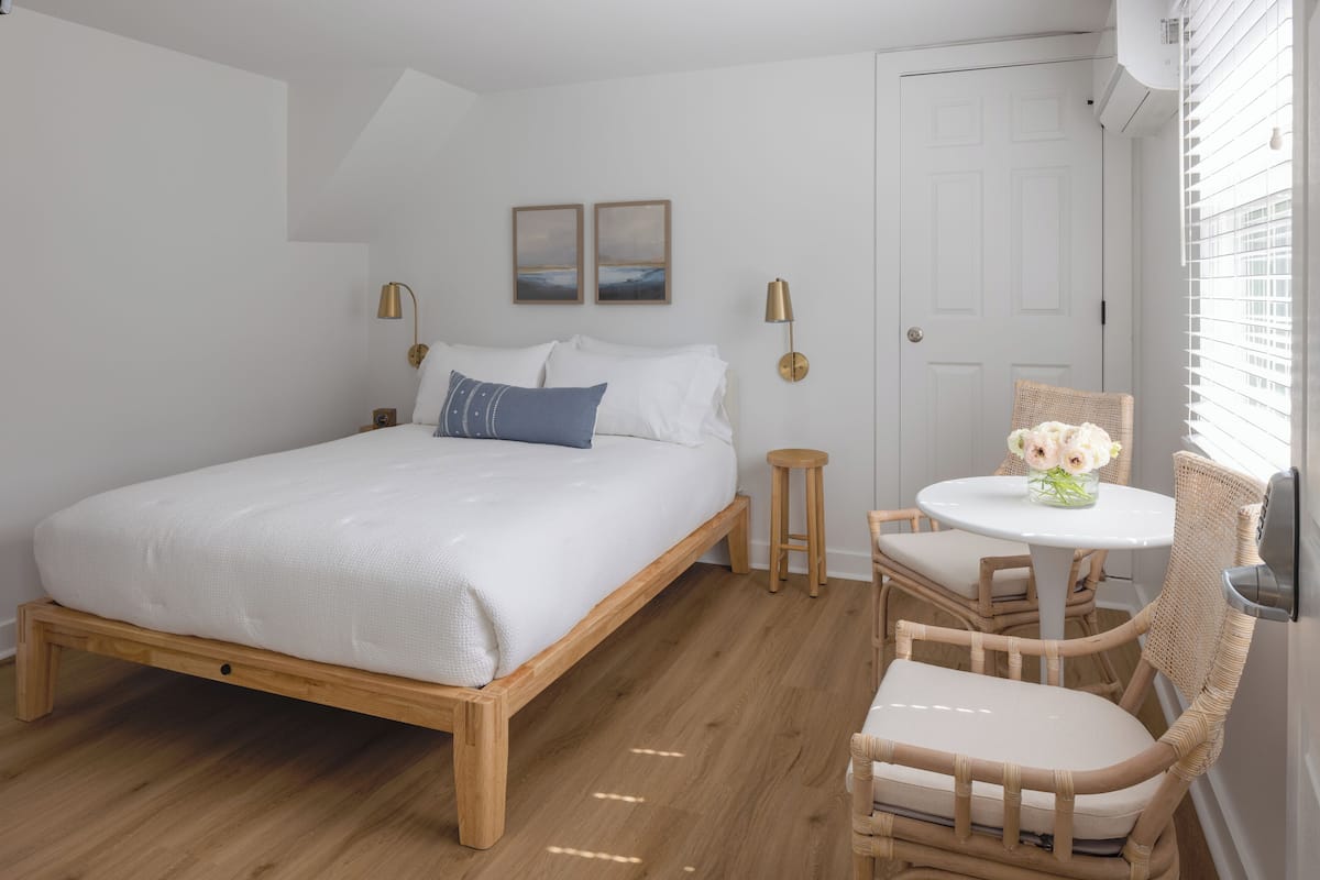 A bedroom with a wooden bed, white bedding, a small round table with chairs, and a wall-mounted air conditioner.
