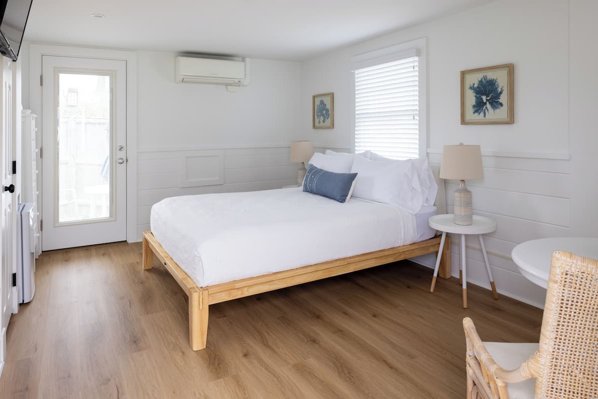 A bedroom with a wooden bed, white bedding, a bedside table with a lamp, and a chair.